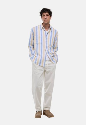 Homme les mains dans les poches, portant une chemise rayée bleue, jaune et blanche, un pantalon blanc et des chaussures marron sans lacets, debout sur un fond blanc.