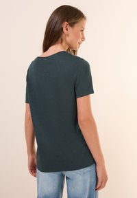 T-shirt corto color verde scuro con scollo rotondo, caratterizzato da una texture liscia e una vestibilità rilassata, abbinato a jeans azzurri chiari.