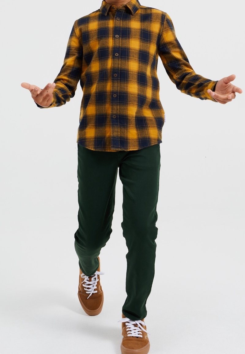 Camicia a quadri gialla e blu navy con bottoni, pantaloni verdi e sneakers marroni con lacci bianchi. La camicia ha un colletto e una chiusura frontale con bottoni.