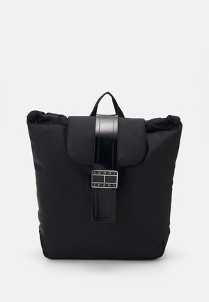 Mochila negra con asa superior, cierre con solapa y una correa brillante con una hebilla rectangular con el logo de Tommy Jeans.