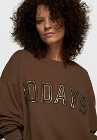 Mujer con cabello rizado que lleva una sudadera marrón con la inscripción "100 DÍAS" en letras tachonadas en el frente, mirando a la cámara con confianza.