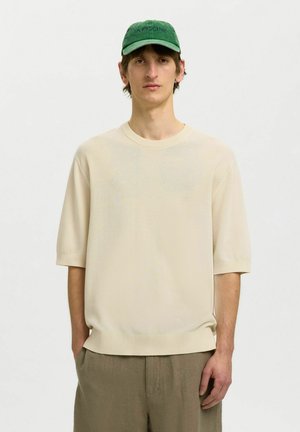 Jeune homme portant un pull court texturé couleur crème, un pantalon vert olive et une casquette de baseball verte avec un texte. Mains dans les poches.