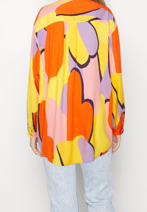 Koszula z długim rękawem z abstrakcyjnymi blokami kolorów w pomarańczowym, żółtym, różowym i fioletowym, noszona z jasnoniebieskimi dżinsami, widok od tyłu.