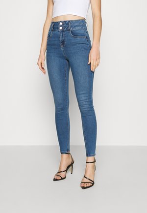 Jean slim fit bleu taille haute avec fermeture à double bouton porté par une personne portant également un crop top blanc et des talons hauts noirs à brides.