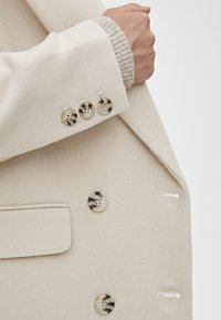 Cappotto in misto lana beige con tre bottoni decorati sul polsino, dotato di un bordo interno a maglia tessuta e una tasca.