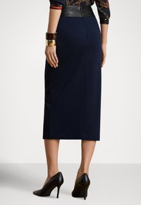 Sieviete no aizmugures, tērpusies tumši zili midi zīmuļsvārkos, ar rakstainu krekliņu ielocītu biksēs, melnas augstpapēžu kurpes un zelta un bruņurupuču krāsas mas�īvas rokassprādzes.