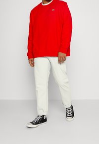 Czerwony sweatshirt, biały okrągły dekolt, luźny krój. Jasnoszare spodnie, lekko podwinięte mankiety. Czarne sneakersy za kostkę z białymi akcentami.