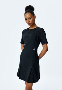 Zwart kortemouwen jurk met een ronde halslijn, slim-fit lijfje en een uitlopende rok met plooien en een decoratieve broche op de taille.