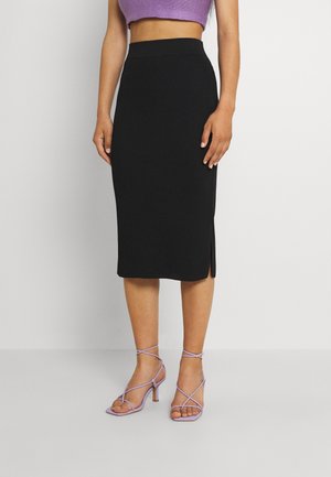 Kvinde iført sort ribbet midi nederdel med sidelommer, lilla cropped top og lavendel stroppesandaler med høje hæle, stående mod en ensfarvet baggrund.