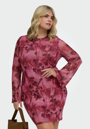 Femme portant une robe rouge à fleurs à manches longues, tenant un sac à main marron, avec des cheveux blonds ondulés et la main sur la hanche, regardant sur le côté.