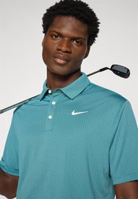Τιρκουάζ polo μπλούζα γκολφ της Nike με διακριτικό υφασμάτινο σχέδιο, τρία κουμπιά και λευκό λογότυπο. Γκολφιέρης ποζάρει με ένα μπαστούνι στους ώμους.