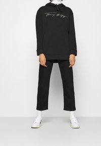 Černá mikina s kapucí na stažení a vyšitým zlatým logem, doplněná černými kalhotami rovného střihu a bílými teniskami.