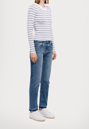 Frau trägt ein weiß-grau gestreiftes Langarmshirt, blaue Jeans und weiße Sneakers und steht vor einem schlichten Hintergrund.