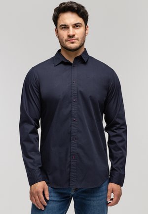 Granatowa koszula z długim rękawem zapinana na guziki, z delikatnym połyskiem, z kontrastowymi czerwonymi guzikami oraz małym haftowanym logo na piersi.