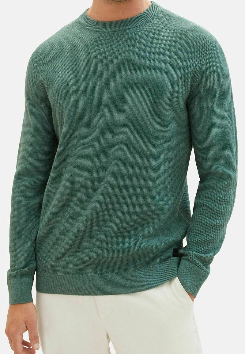 Homme portant un pull vert texturé à manches longues avec col rond et un pantalon blanc, une main dans la poche, sur un fond uni.