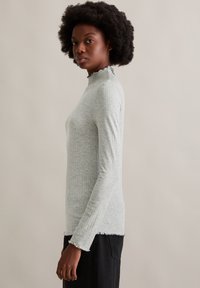 Graues Langarm-Oberteil mit gerippter Textur und einem frillierten Stehkragen, mit rohen Kanten. Getragen mit schwarzen Hosen.