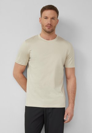 Mann mit kurzen braunen Haaren, der ein beiges kurzärmeliges T-Shirt und schwarze Hosen trägt, steht vor einer hellgrauen Wand.