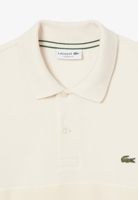 Polo beige en coton texturé, avec un plastron à deux boutons, un col côtelé avec des rayures vertes et un logo de crocodile vert.