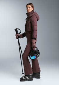 Ski-outfit met een donkergroen gewatteerd jack met capuchon, bijpassende nauwsluitende broek, zwarte skischoenen, skistokken en een helm met een blauw vizier.