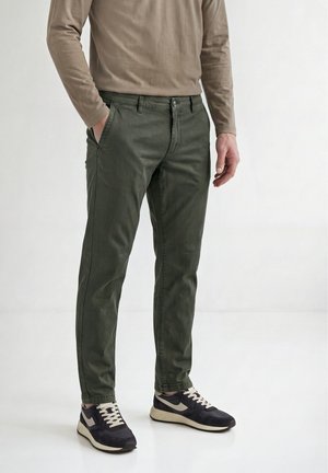 Mann trägt olivgrüne Hose, beige Langarmshirt und schwarz-weiße Sneaker, steht mit einer Hand in der Tasche.