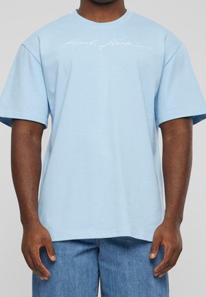 Mand iført en lyseblå oversized T-shirt med subtil hvid skrift over brystet og blå jeans, stående med afslappede arme.