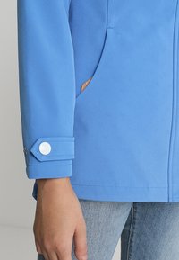 Mano en el bolsillo de una chaqueta azul claro con puño blanco abotonado, combinada con jeans de mezclilla azul claro, sobre un fondo neutro.