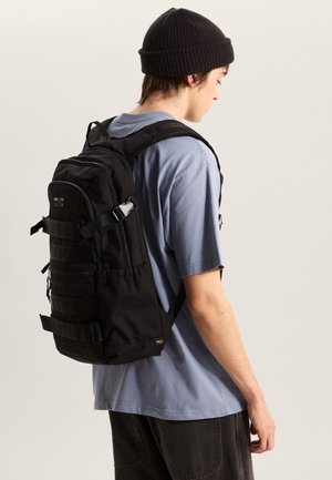 Mochila negra hecha de tela resistente con múltiples compartimentos, correas ajustables y una superficie texturizada. Se lleva en la espalda de una persona.