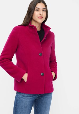 Frau trägt einen knallpinkfarbenen, geknöpften Mantel mit Händen in den Taschen und blaue Jeans, steht vor einem weißen Hintergrund.