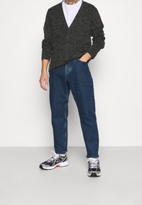 Man som bär en mörkgrå knappad cardigan över vit skjorta, blå denimjeans och svart-grå sneakers står framför en vit bakgrund.