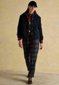 Giacca in pile navy con dettagli marroni, maglione rosso, pantaloni in plaid verde e scarpe scure. Tessuto testurizzato, dettagli con bottoni e tasche visibili.