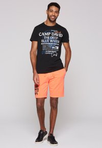 Svart t-shirt med grafiskt tryck, kombinerad med ljus orange shorts som har en logotyp. Modell som bär svarta sneakers.