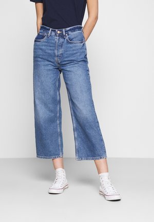 Vaaleansinisiä korkea vyötäröisiä leveitä farkkuja, joissa on etutaskut ja näkyvät saumat. Yhdistettynä valkoisiin korkeisiin tennareihin.