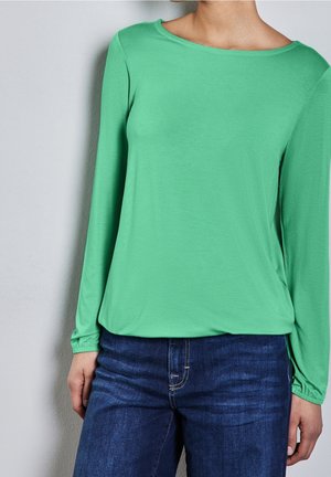 Vrouw draagt een groen top met lange mouwen in combinatie met donkerblauwe spijkerbroek, staand tegen een effen lichte muur.