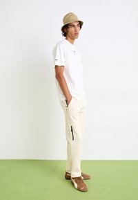 White cotton t-shirt, fitted, with Puma logo. Beige utility bucket hat. Cream tapered joggers with zip pockets and black accents, brown sneakers.