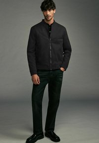 Schwarze Bomberjacke mit Reißverschluss, gerippten Bündchen und glatter Oberfläche, kombiniert mit dunklen Jeans und glänzenden schwarzen Schuhen.