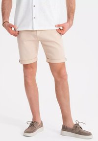 Shorts beige avec des ourlets roulés, fabriqués en tissu de coton. Portés avec une chemise blanche à boutons et des chaussures en daim gris. Texture lisse.
