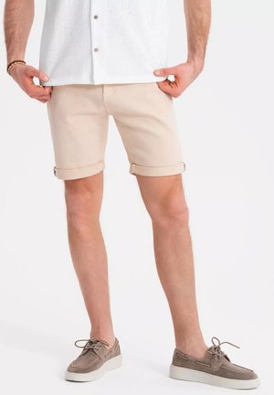 Pantaloni corti beige con risvolti, realizzati in tessuto di cotone. Indossati con una camicia bianca a maniche lunghe e scarpe in suede grigio. Tessura liscia.