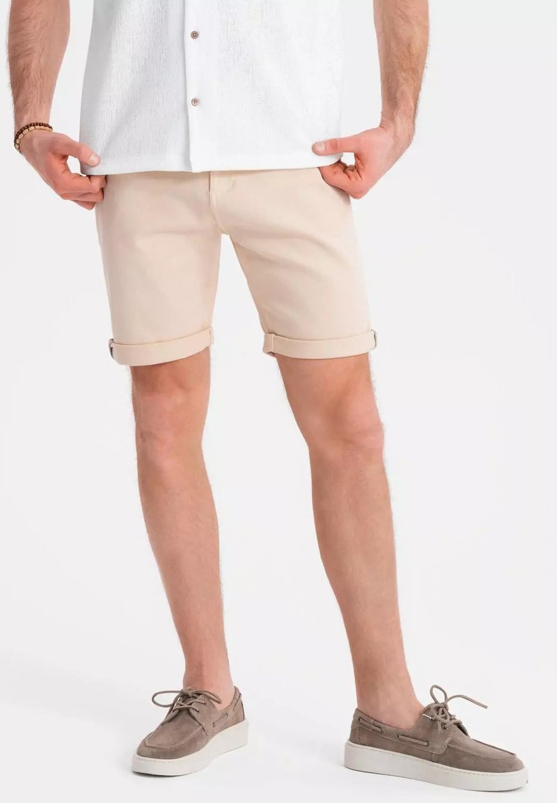 Beige shorts dengan lipatan, terbuat dari kain katun. Dipadukan dengan kemeja putih berkerah dan sepatu suede abu-abu. Tekstur yang halus.