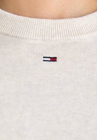 Pull en tricot beige avec un col côtelé, comportant un petit logo de drapeau brodé en rouge, blanc et bleu près du col.