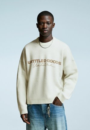 Junger Mann trägt einen cremefarbenen Pullover mit dem Text "Untitled Goods", blaue Jeans, eine silberne Kette um den Hals und hat Schlüssel an seiner Tasche befestigt, vor einem schlichten Hintergrund.