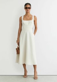 Leveäolkaiminen, suorakauluksinen mekko off-white-värissä, jossa on istuva yläosa ja A-linjainen helma, sivutaskuilla. Ruskea olkalaukku ja remmikkäät korkokengät.