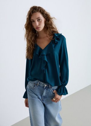 Donna con capelli lunghi e mossi indossa una blusa verde acqua con volant e jeans azzurri, in piedi davanti a uno sfondo bianco e grigio semplice.