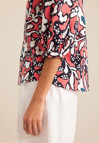 Bluzka w kwiatowy wzór w kolorze koralowym, granatowym i białym, z bufkami i zaokrąglonym dołem. Połączona z jasnymi spodniami lnianymi.