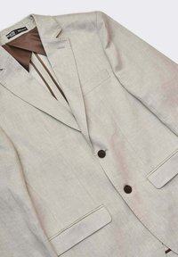 Ljust beige blazer i linneblandning med snedslåkrage, två framfickor och bruna knappar. Inuti har den brun foder med ränder.
