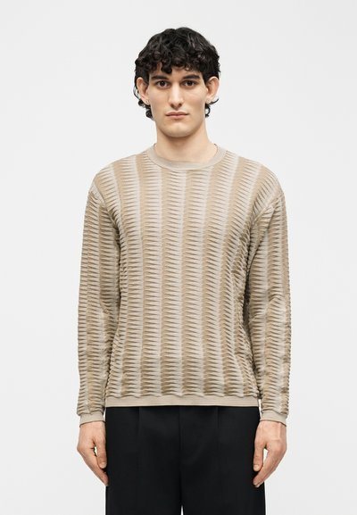 Homme aux cheveux courts et bouclés portant une chemise beige texturée à manches longues et un pantalon noir, debout devant un fond clair uni.