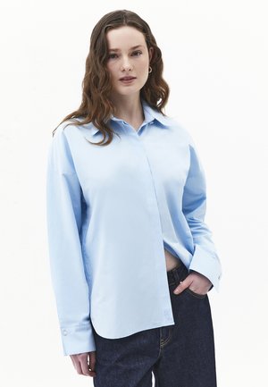 Młoda kobieta o falowanych brązowych włosach, ubrana w jasnoniebieską, luźną koszulę i ciemne jeansy, stoi z jedną ręką w kieszeni na białym tle.