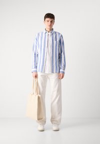 Gestreept blauw en wit button-up shirt, losse witte broek en een beige tote bag. De outfit heeft casual sneakers en eenvoudige lijnen.