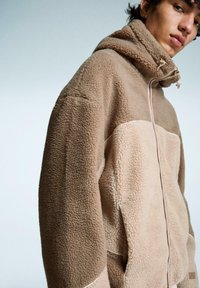 Jachetă din fleece maro cu fermoar frontal, guler înalt și o pată contrastantă mai deschisă. Textură moale și glugă cu șnur ajustabil.