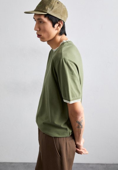 T-shirt verde oliva a maniche corte con cuciture a contrasto bianche, abbinato a pantaloni marroni. Il modello indossa un cappellino beige. Tatuaggio visibile sul braccio.