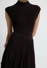 Černé žebrované šaty s vysokým výstřihem a krátkými rukávy, které mají upnutou korzetovou část a splývavou, plisovanou sukni. Je viditelný texturovaný detail látky.